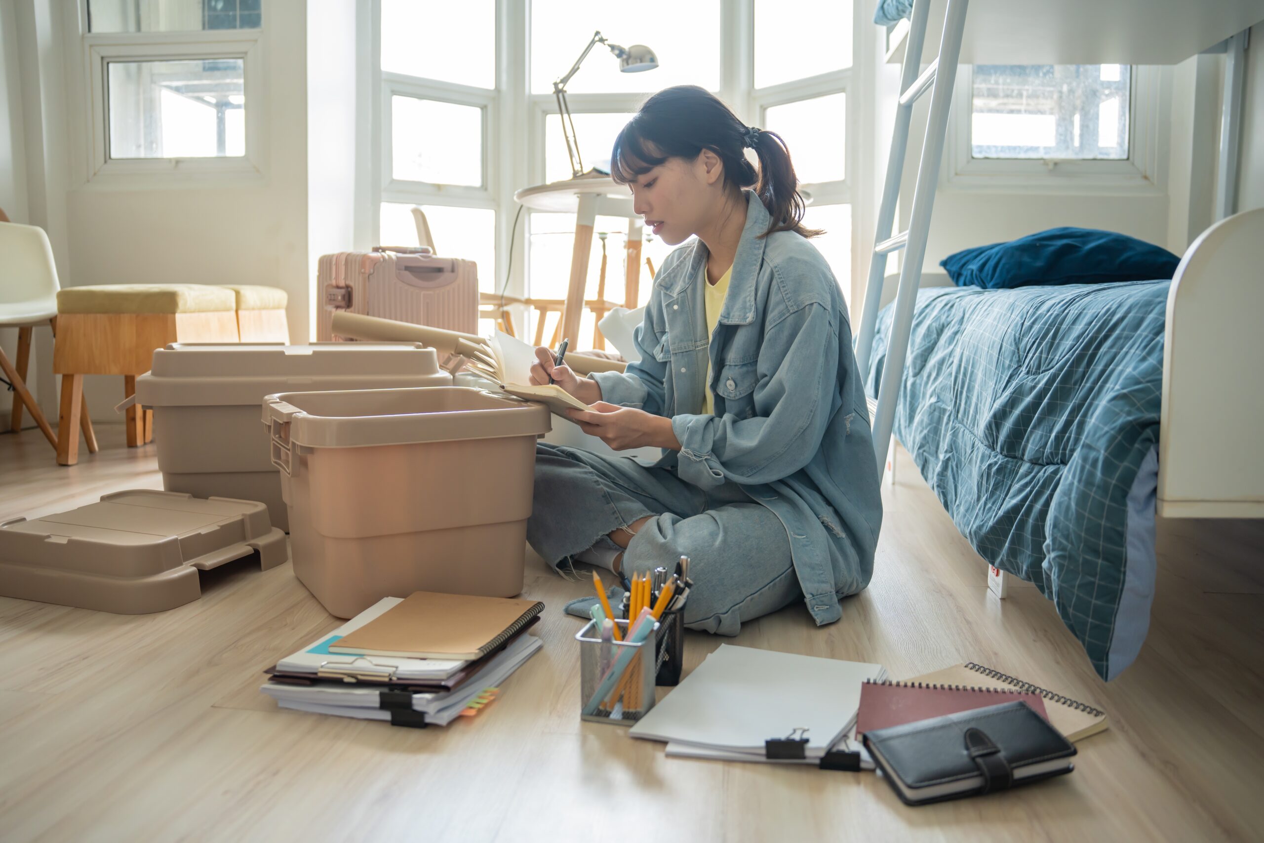 Female student writing in a notepad in dorm room with plastic tubs and luggages