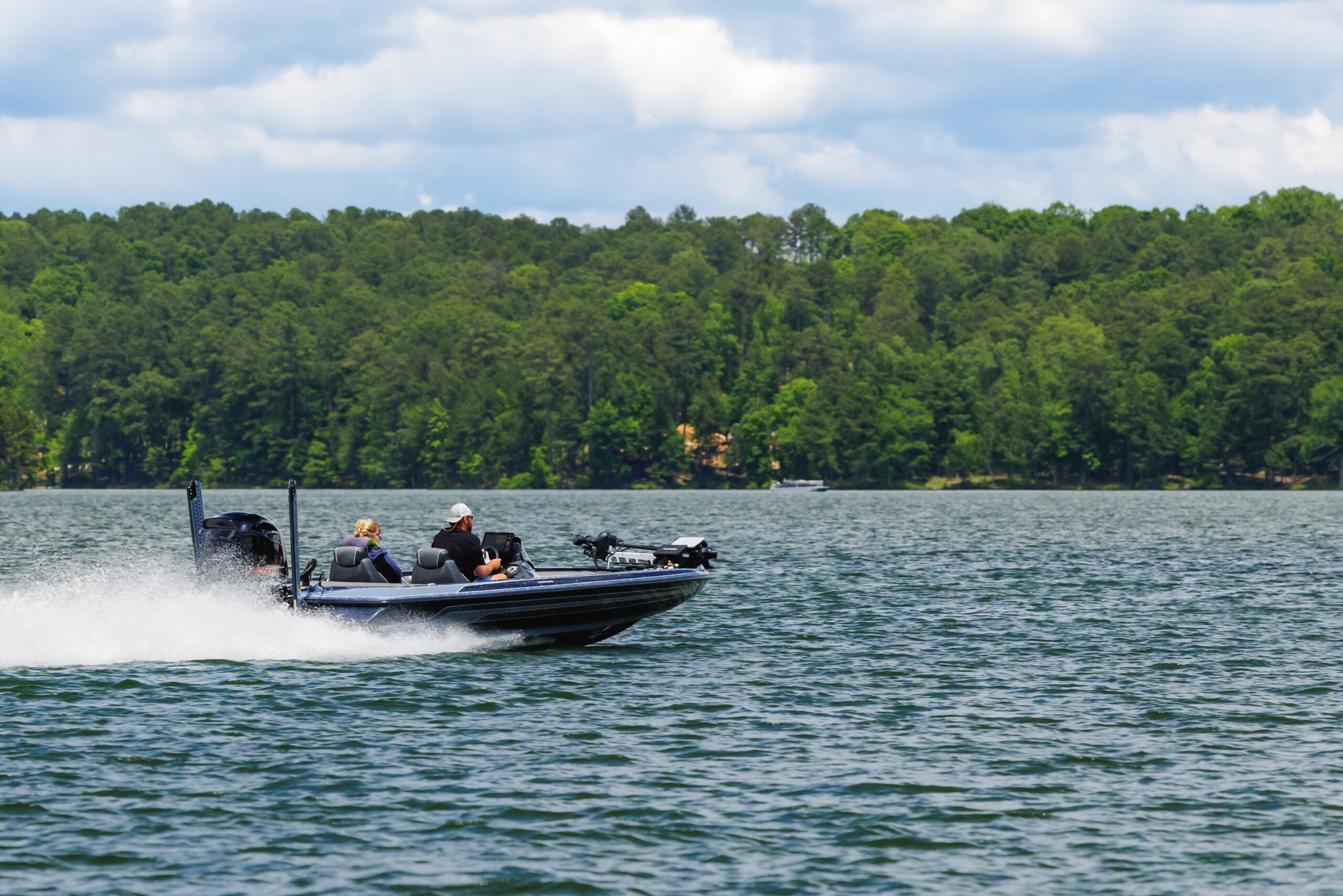 A bass fishing boat with two people speeds across a large lake, kicking up spray, illustrating the specialized nature of different recreational types of boats. A dense forest line is in the background.