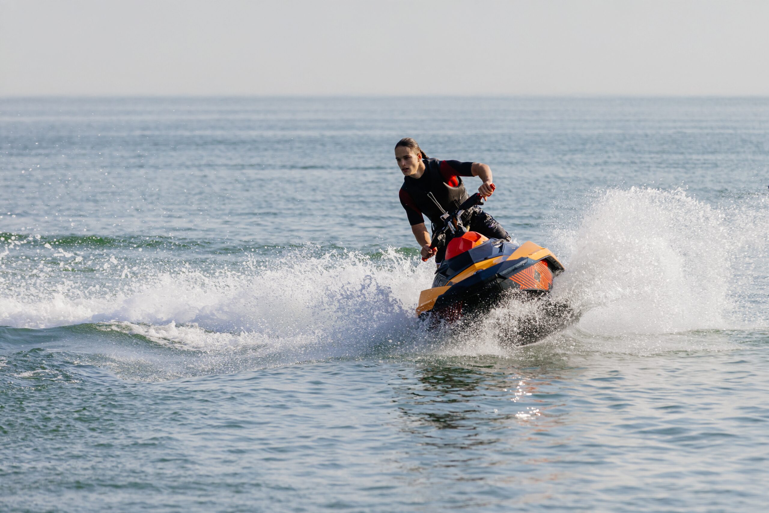 A person in a life jacket and wetsuit powerfully turns an orange and blue personal watercraft (PWC), creating a big splash in the open ocean, showcasing a thrilling and agile example within the spectrum of available types of boats.