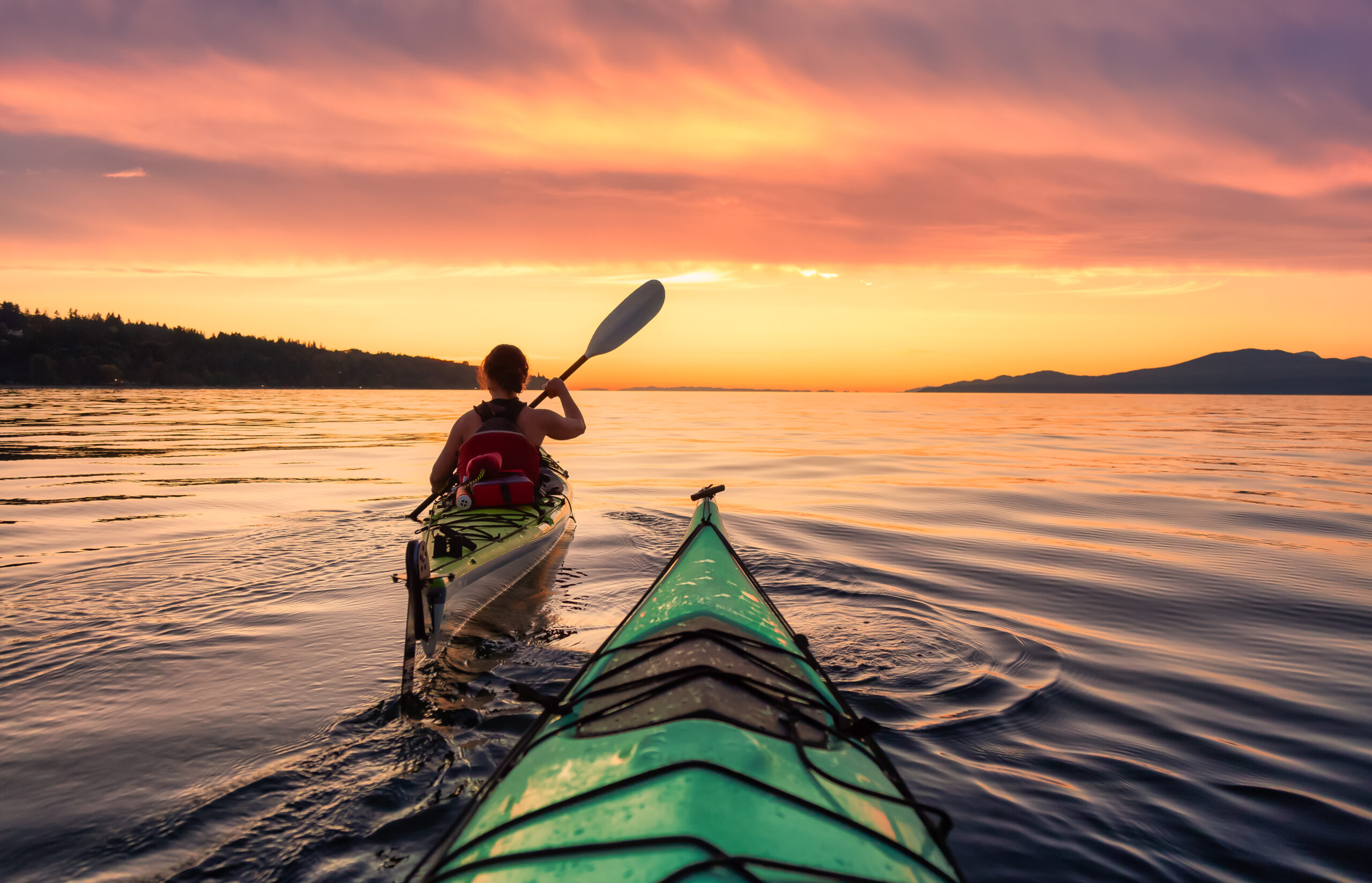 First-person view of a green and yellow sea kayak paddling on a calm ocean at sunset, with a person just ahead in their own kayak, highlighting human-powered options for the different recreational types of boats.