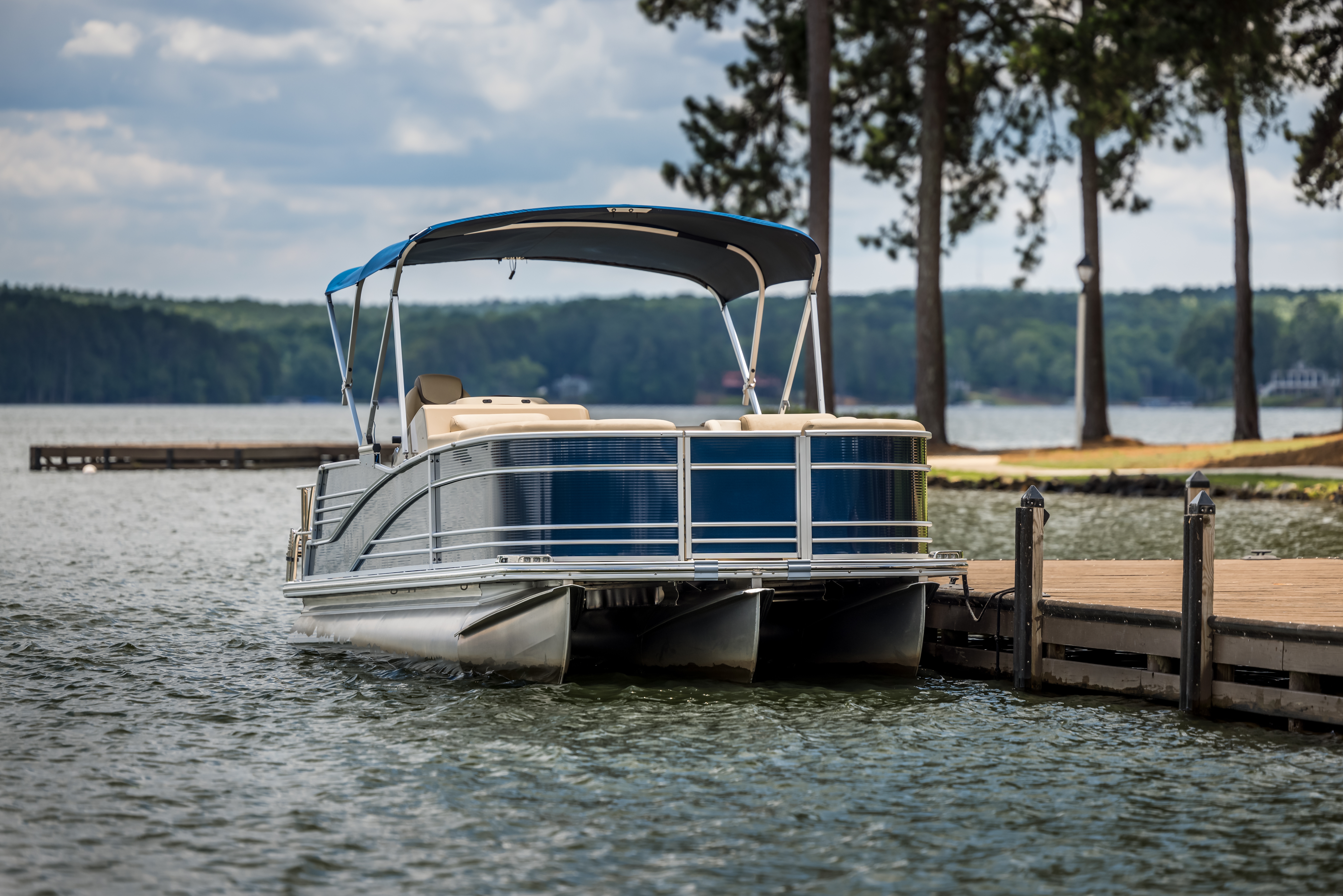 A blue and beige pontoon boat with a bimini top is securely docked at a wooden pier on a lake, with pine trees on the opposite shore, showing a stable and social layout found in common types of boats.