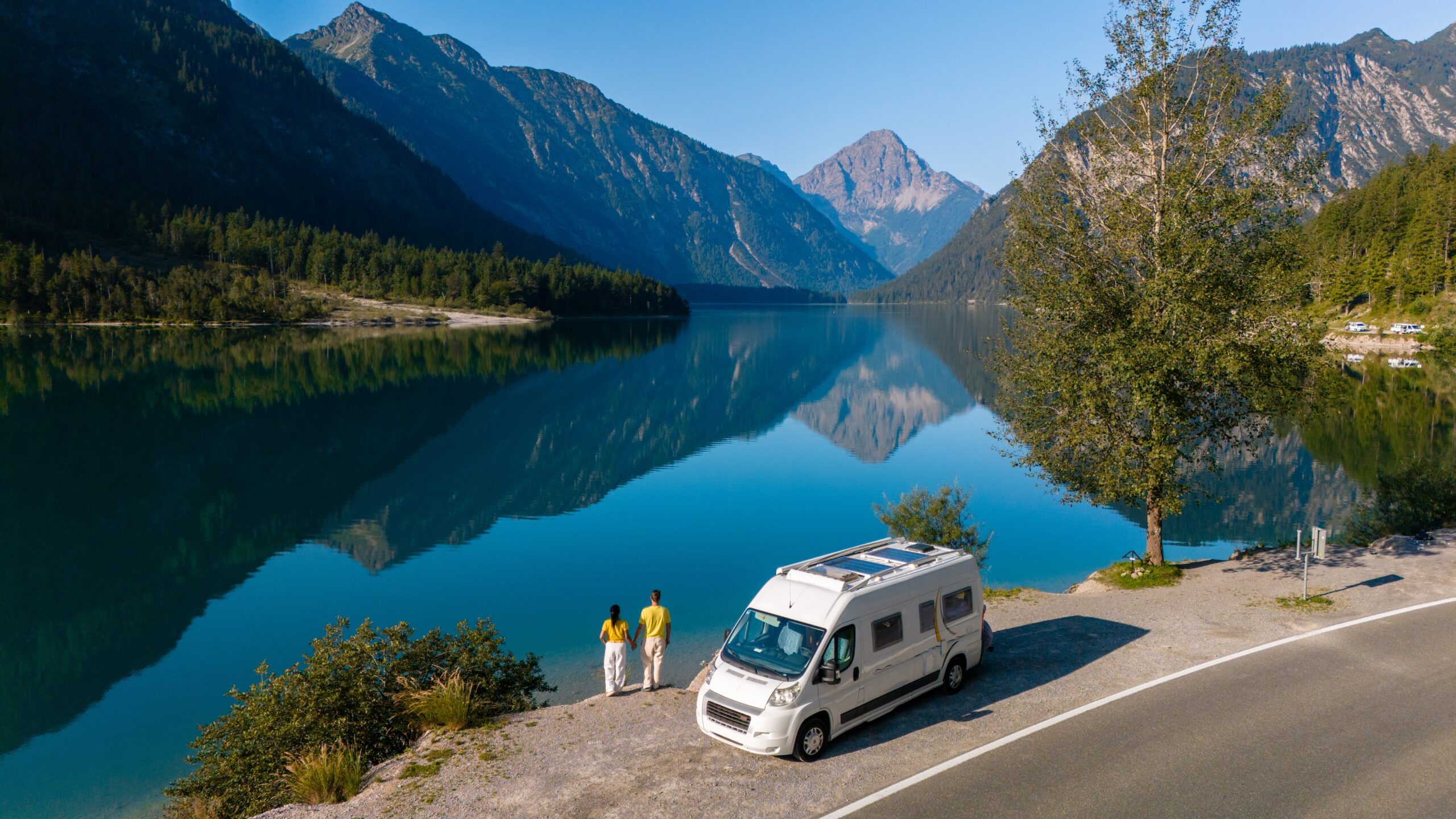 A high-angle aerial photograph of a compact white camper van parked on a scenic roadside next to a glassy alpine lake that reflects large mountains. A couple stands hand-in-hand at the water's edge next to the vehicle, showcasing a smaller Class B choice within typical average RV sizes.